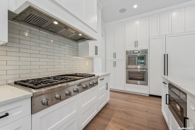 kitchen featuring light stone countertops, premium range hood, tasteful backsplash, built in appliances, and white cabinetry