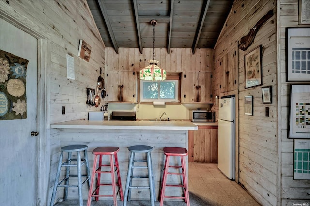 kitchen with vaulted ceiling with beams, kitchen peninsula, wood walls, white fridge, and pendant lighting