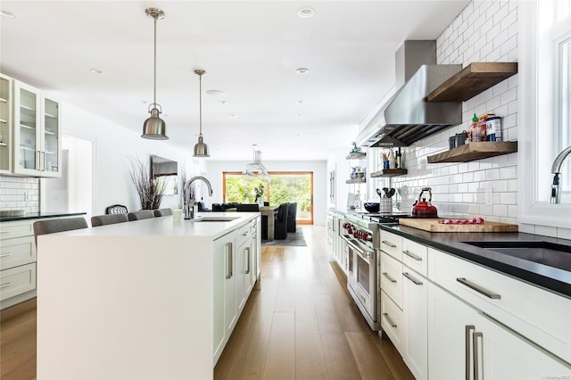 kitchen with wall chimney range hood, pendant lighting, double oven range, a kitchen island with sink, and white cabinets