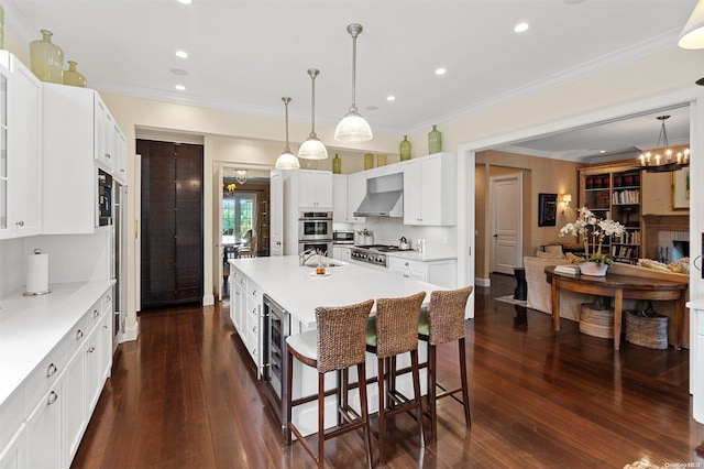 kitchen featuring dark wood-type flooring, a center island with sink, a kitchen breakfast bar, wall chimney range hood, and white cabinetry