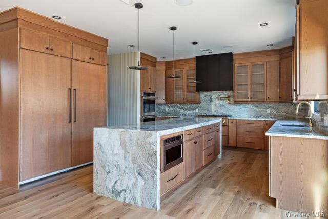 kitchen featuring stainless steel microwave, light wood-type flooring, light stone counters, exhaust hood, and a sink