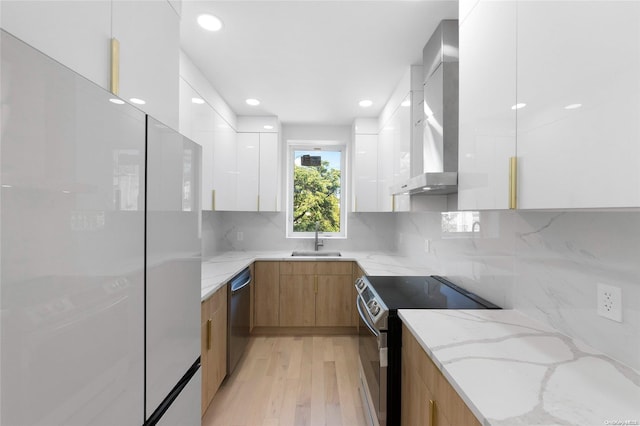 kitchen featuring light stone counters, wall chimney exhaust hood, stainless steel appliances, sink, and white cabinetry