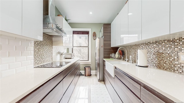 kitchen featuring wall chimney exhaust hood, stainless steel fridge, black electric cooktop, tasteful backsplash, and white cabinetry