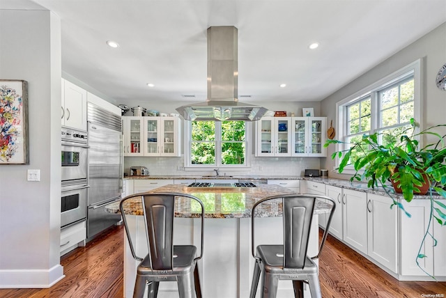 kitchen featuring light stone countertops, a kitchen breakfast bar, island range hood, hardwood / wood-style flooring, and white cabinets