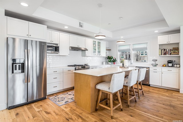 kitchen with a kitchen island, a raised ceiling, appliances with stainless steel finishes, white cabinets, and light wood-type flooring