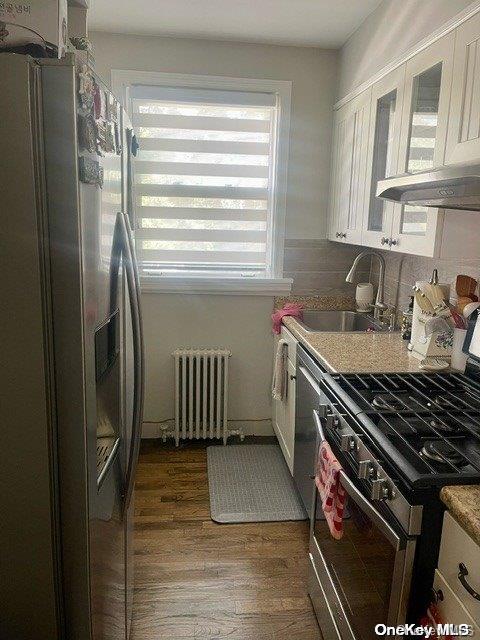 kitchen with radiator, sink, appliances with stainless steel finishes, white cabinets, and hardwood / wood-style flooring