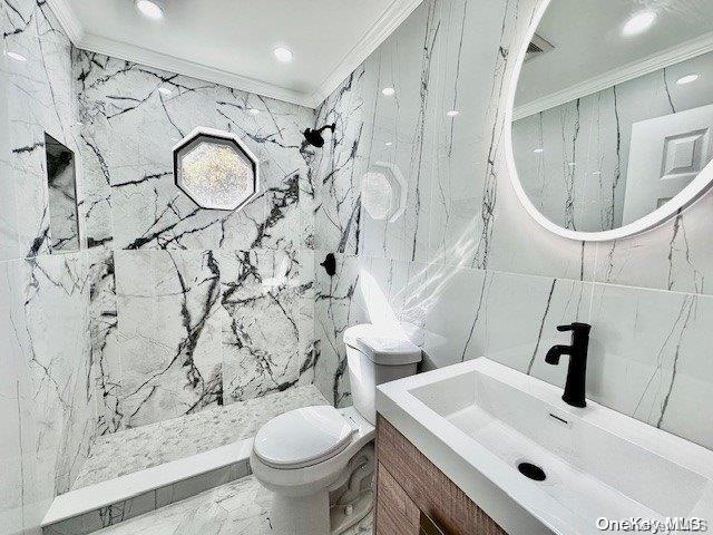 bathroom featuring a tile shower, crown molding, toilet, vanity, and tile walls