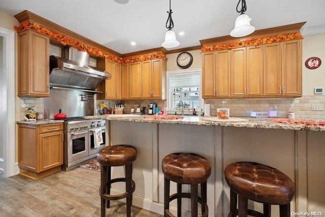 kitchen featuring wall chimney exhaust hood, light stone countertops, appliances with stainless steel finishes, decorative light fixtures, and a kitchen bar