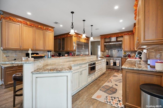 kitchen featuring wall chimney range hood, high quality appliances, light hardwood / wood-style floors, a kitchen bar, and decorative backsplash