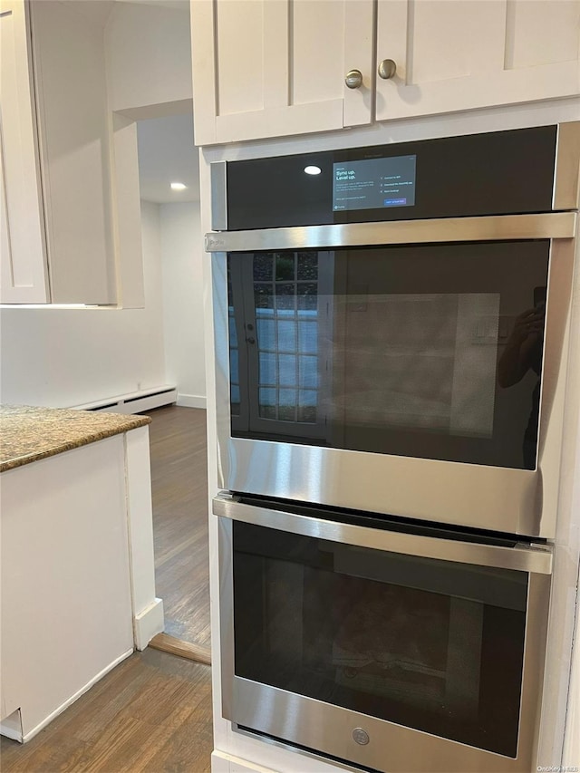 room details featuring stainless steel double oven, dark wood-type flooring, light stone counters, a baseboard heating unit, and white cabinets