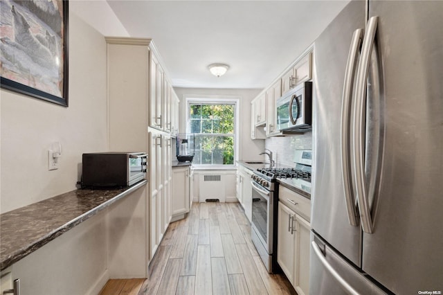 kitchen featuring appliances with stainless steel finishes, light wood-type flooring, radiator, dark stone counters, and white cabinets