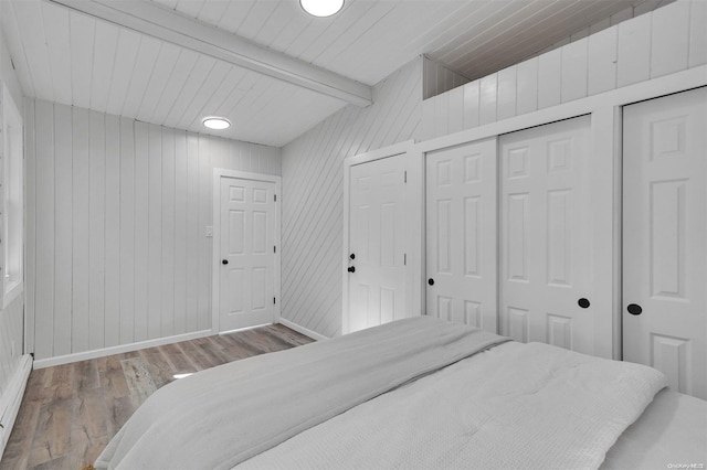 bedroom with two closets, baseboard heating, wooden walls, beam ceiling, and hardwood / wood-style flooring