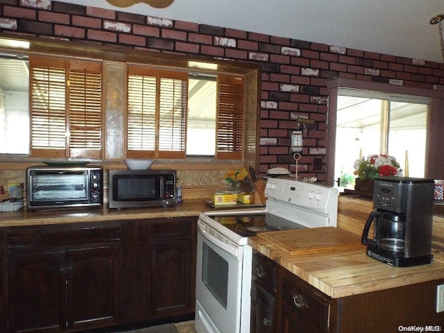 kitchen featuring white range with electric stovetop, dark brown cabinetry, a healthy amount of sunlight, and brick wall