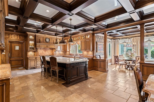 kitchen featuring a center island with sink, decorative light fixtures, coffered ceiling, and decorative columns