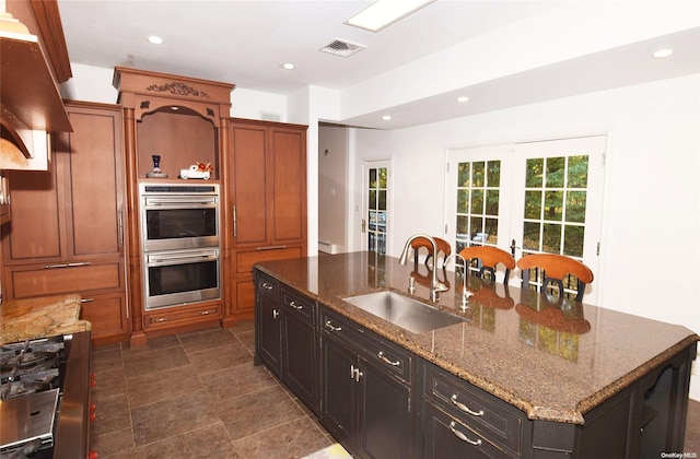 kitchen featuring dark stone counters, french doors, sink, an island with sink, and stainless steel double oven