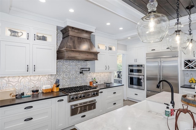 kitchen featuring decorative backsplash, custom range hood, stainless steel appliances, white cabinets, and hanging light fixtures