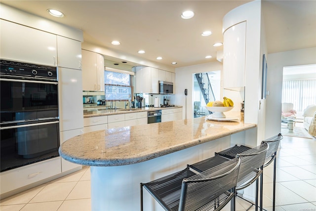 kitchen featuring white cabinetry, light stone counters, double oven, a breakfast bar, and light tile patterned floors