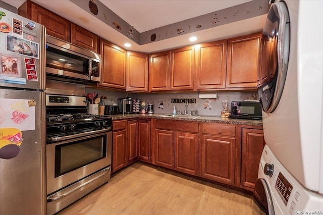 kitchen with sink, dark stone countertops, light hardwood / wood-style floors, stacked washer / dryer, and stainless steel appliances