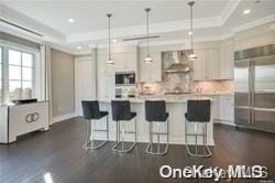 kitchen with high end refrigerator, tasteful backsplash, a raised ceiling, wall chimney range hood, and decorative light fixtures