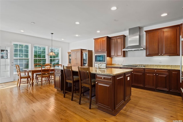 kitchen with wall chimney exhaust hood, sink, pendant lighting, a center island with sink, and light hardwood / wood-style floors