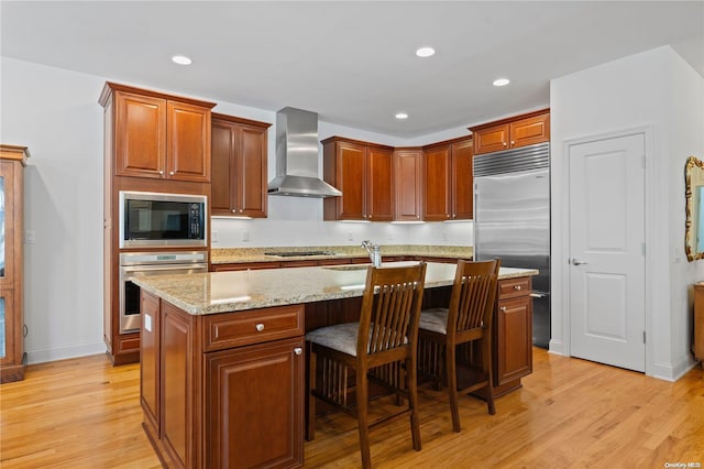 kitchen featuring wall chimney range hood, built in appliances, light hardwood / wood-style floors, a breakfast bar area, and a kitchen island with sink