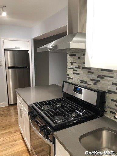 kitchen featuring white cabinetry, wall chimney exhaust hood, stainless steel appliances, tasteful backsplash, and light wood-type flooring