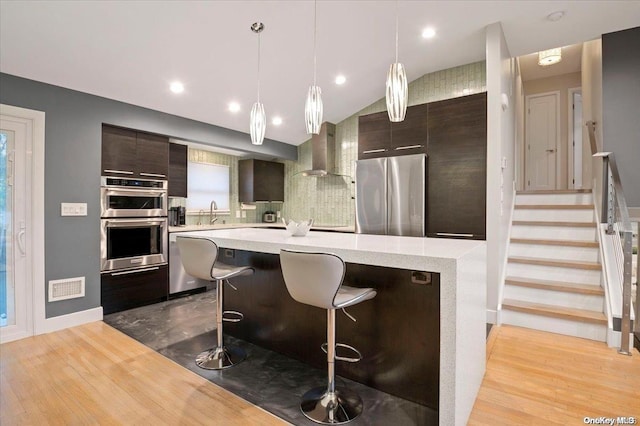 kitchen featuring a kitchen breakfast bar, light wood-type flooring, dark brown cabinets, stainless steel appliances, and wall chimney range hood