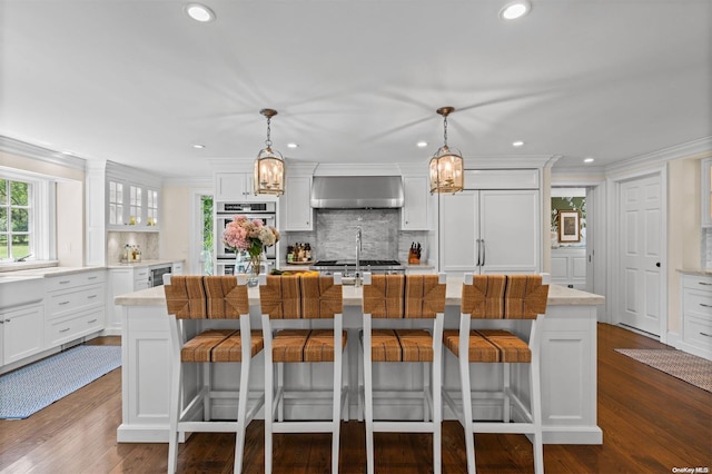 kitchen with wall chimney exhaust hood, stainless steel double oven, dark wood-type flooring, hanging light fixtures, and an island with sink