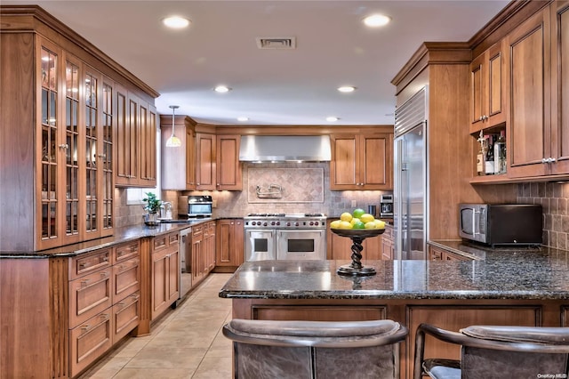 kitchen with hanging light fixtures, wall chimney range hood, dark stone counters, a breakfast bar area, and high end appliances