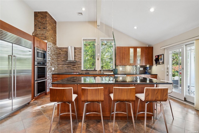 kitchen featuring wall chimney exhaust hood, tasteful backsplash, built in appliances, a breakfast bar area, and a kitchen island