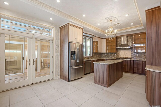 kitchen featuring a center island, a raised ceiling, wall chimney range hood, appliances with stainless steel finishes, and decorative light fixtures