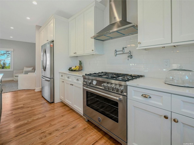 kitchen with appliances with stainless steel finishes, white cabinetry, backsplash, light hardwood / wood-style floors, and wall chimney range hood