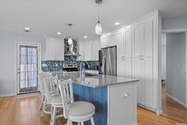 kitchen featuring white cabinetry, wall chimney exhaust hood, stainless steel appliances, light hardwood / wood-style flooring, and an island with sink