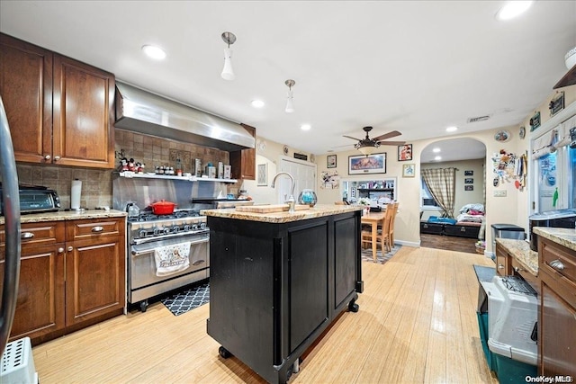 kitchen with light wood-type flooring, backsplash, wall chimney range hood, high end stainless steel range, and an island with sink