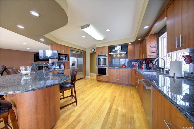 kitchen featuring a breakfast bar, built in appliances, wall chimney range hood, and sink