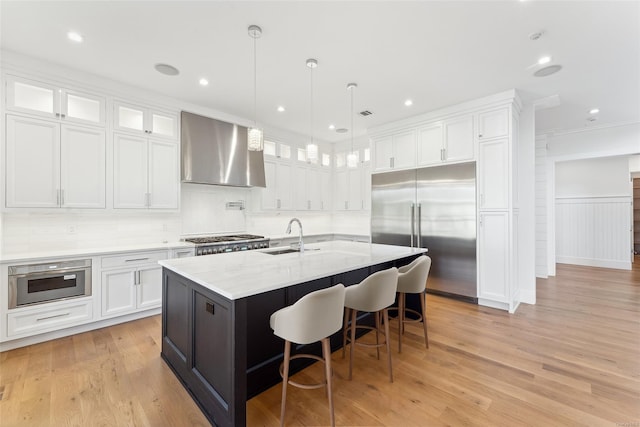 kitchen featuring wall chimney exhaust hood, stainless steel appliances, a center island with sink, light hardwood / wood-style flooring, and white cabinetry