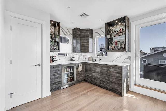 bar featuring light wood-type flooring, tasteful backsplash, dark brown cabinetry, sink, and wine cooler