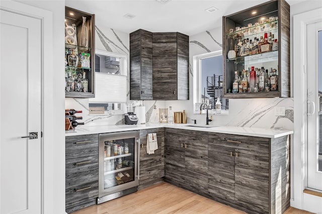 bar featuring sink, light hardwood / wood-style flooring, tasteful backsplash, dark brown cabinetry, and beverage cooler