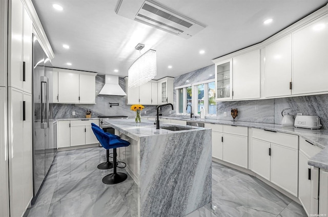 kitchen with white cabinetry, light stone countertops, custom range hood, and a kitchen island with sink