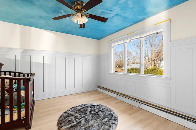 bedroom with ceiling fan, light wood-type flooring, a nursery area, and a baseboard heating unit