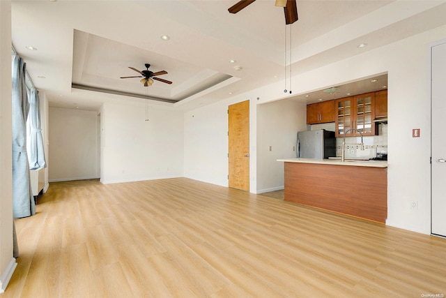 unfurnished living room with ceiling fan, a raised ceiling, sink, and light hardwood / wood-style flooring