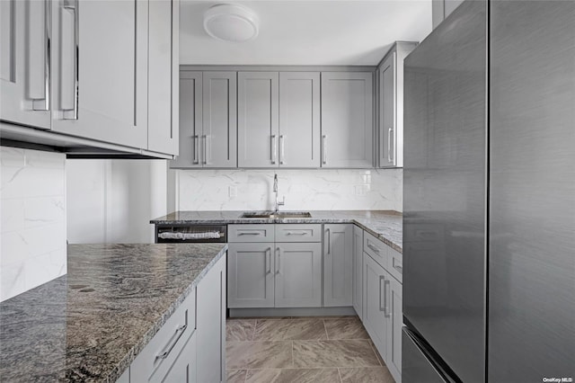 kitchen featuring stainless steel fridge, backsplash, gray cabinetry, dark stone counters, and sink