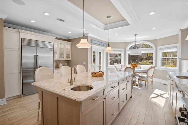 kitchen with a kitchen island with sink, crown molding, sink, built in refrigerator, and light stone countertops