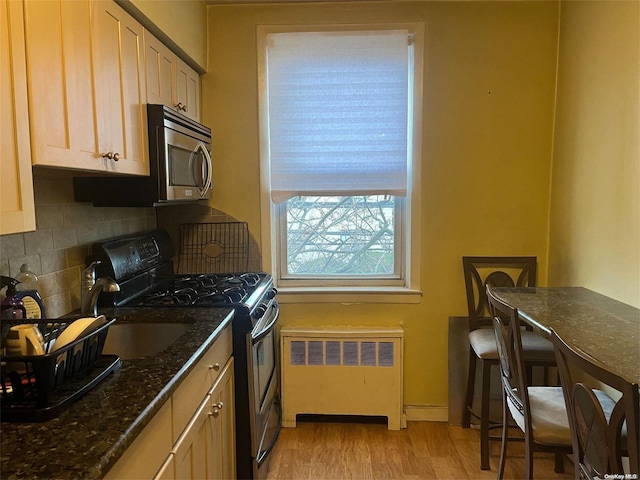 kitchen featuring radiator, dark stone countertops, light brown cabinetry, appliances with stainless steel finishes, and light wood-type flooring