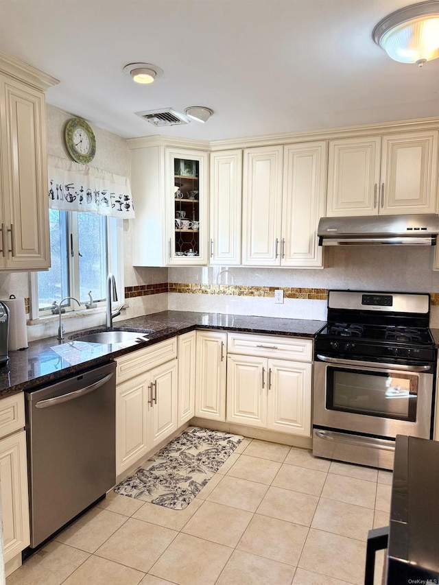 kitchen with dark stone countertops, cream cabinetry, appliances with stainless steel finishes, light tile patterned floors, and sink