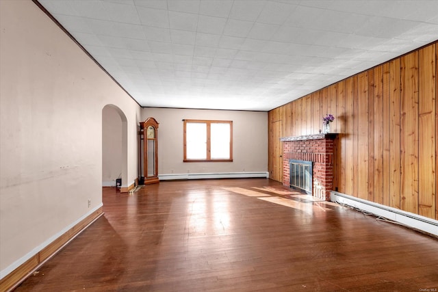 unfurnished living room featuring a brick fireplace, wood-type flooring, wooden walls, and a baseboard radiator