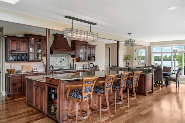 kitchen with custom range hood, light stone counters, a center island with sink, and decorative light fixtures