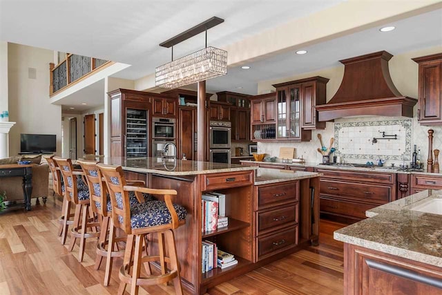 kitchen with decorative light fixtures, light stone counters, tasteful backsplash, a center island with sink, and custom range hood