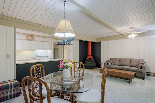 dining area featuring baseboard heating, a wood stove, beam ceiling, ceiling fan, and light colored carpet