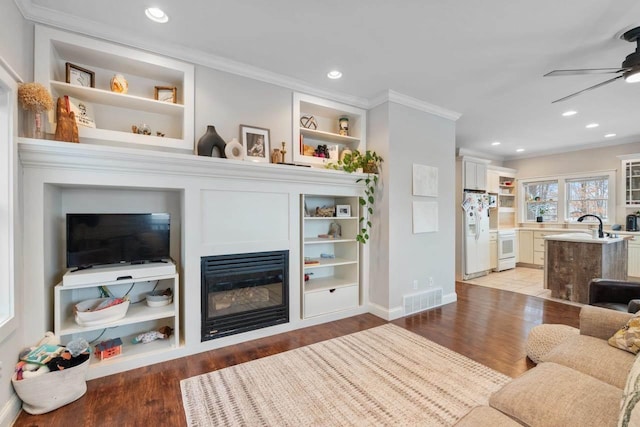 living room featuring crown molding, visible vents, a ceiling fan, a glass covered fireplace, and wood finished floors